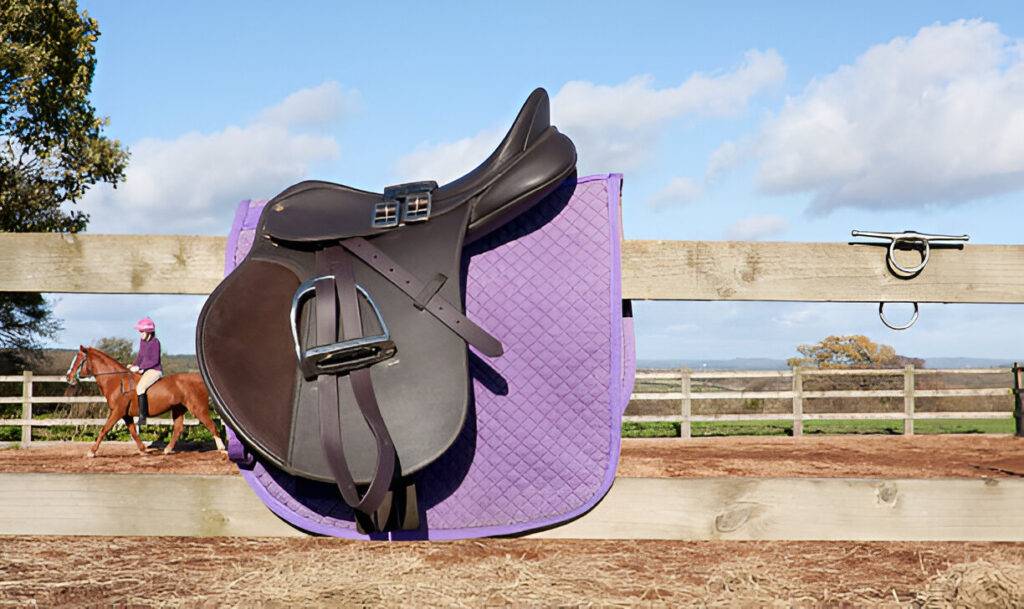 Purple quilted Ogilvy Saddle Pad with a brown English saddle resting on a wooden fence outdoors.
