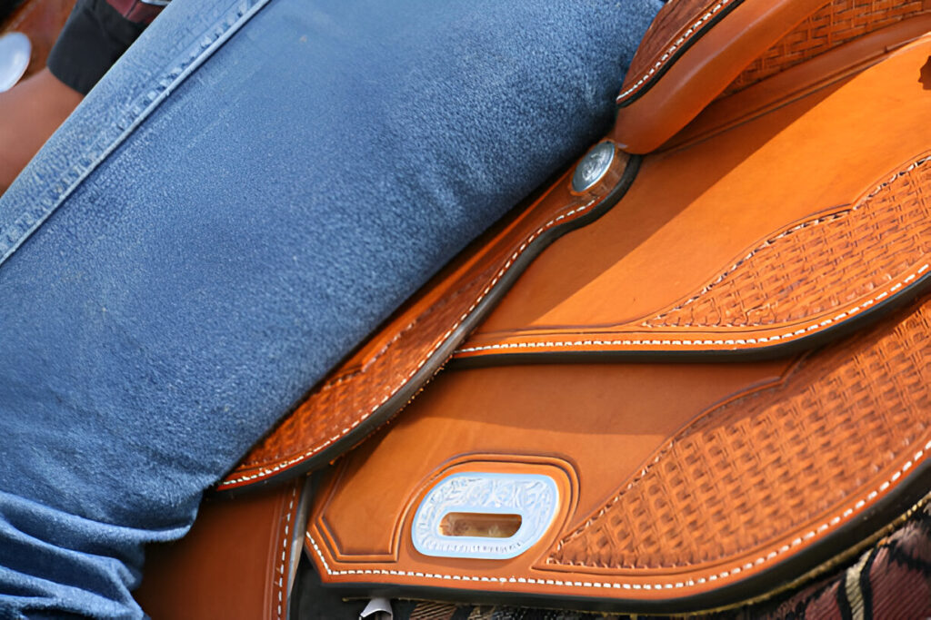 Close-up of an orange Sheridan Saddle with basket‐weave tooling, white stitching, and silver conchos, with a rider’s blue‐jeans–clad leg resting on the fender.
